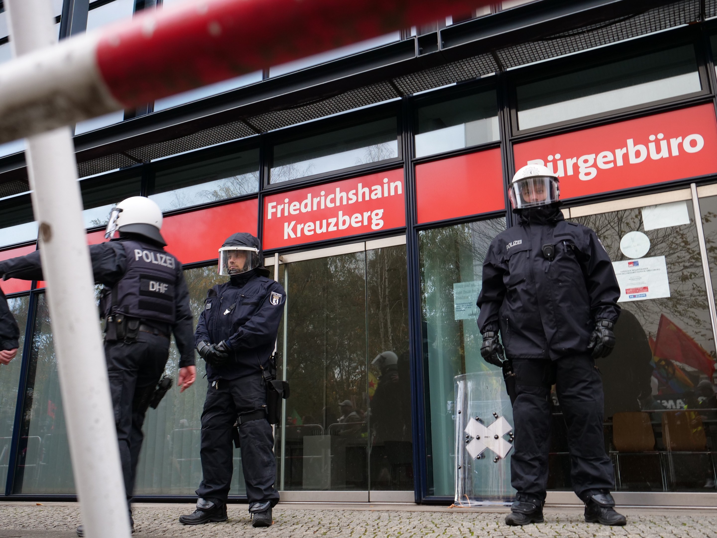 Polizeibeamte in Uniform vor einem Glasfrontgebäude, einige mit Helmen und Handschuhen, mit einer Stange links und Schildern "Friedrichshain Kreuzberg" im Hintergrund.