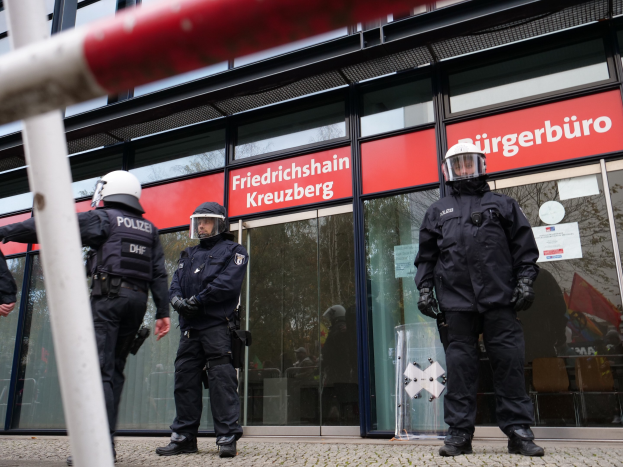 Polizeibeamte in Uniform vor einem Glasfrontgebäude, einige mit Helmen und Handschuhen, mit einer Stange links und Schildern "Friedrichshain Kreuzberg" im Hintergrund.
