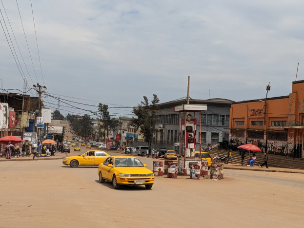 Ein gelbes Taxi fährt eine belebte Stadtstraße mit Häusern, Fußgängern mit Schirmen auf dem Gehweg und Strommasten mit Drähten im Hintergrund unter einem bewölkten Himmel hinunter.