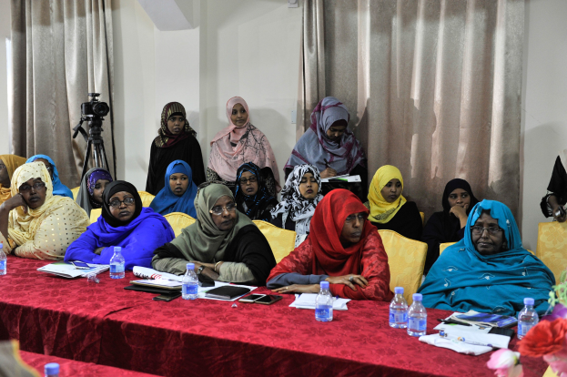 Eine Gruppe von Frauen sitzt an einem Tisch mit verschiedenen Gegenständen, diskutiert engagiert, mit einer Blumenvase und einer Vorhangwand im Hintergrund.