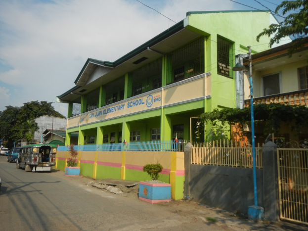 Ein Schulgebäude mit grüner und gelber Fassade an einer Straße, umgeben von Fahrzeugen, Topfpflanzen, einem Zaun, einem Tor, einem Straßenpfahl, Bäumen, Drähten und einem bewölkten Himmel.