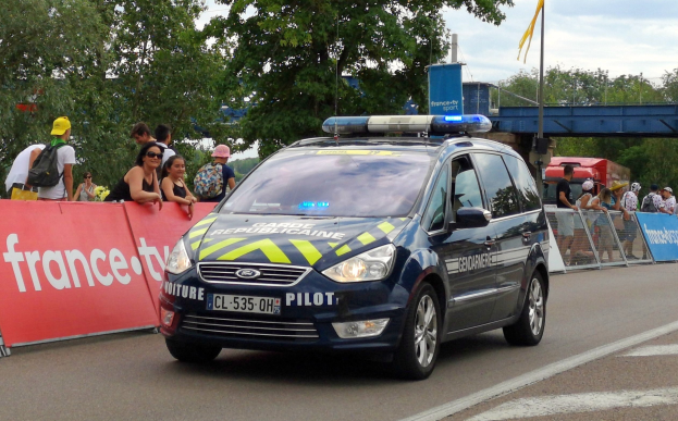Polizeiauto fahrt an einer Menge mit Schildern, Gel├Ąndern, B├Ąumen, einer Brücke, einer Flagge und einem bew├Âlktem Himmel im Hintergrund vorbei.