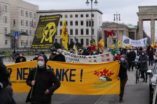 Eine große Gruppe von Menschen marschiert mit Transparenten und Fahnen auf einer Straße bei einer Demonstration gegen Atomkraft in Deutschland, mit Fahrzeugen auf der rechten Seite und Gebäuden, Lötpälen, Bäumen und einem Tor im Hintergrund.
