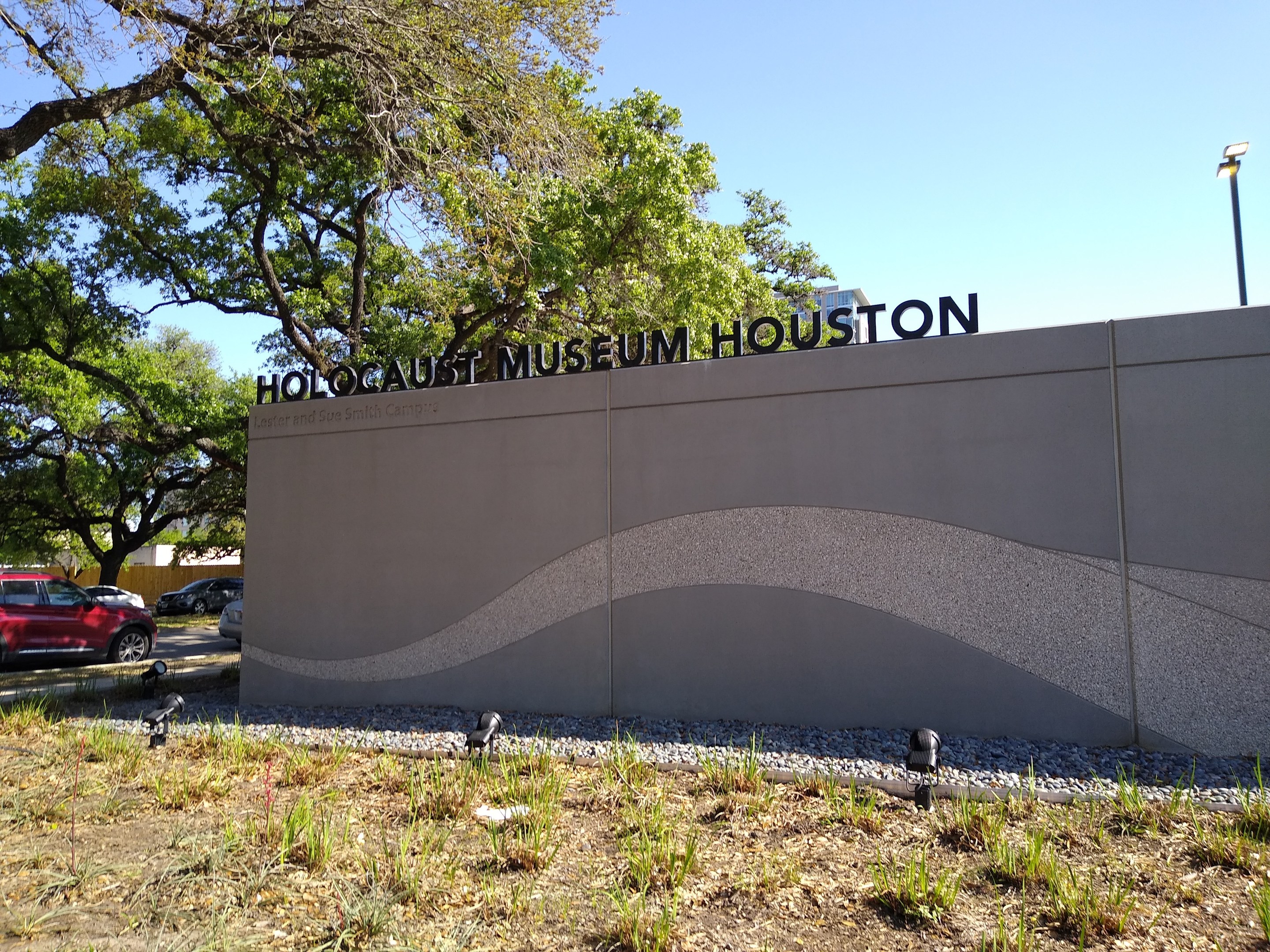 Außenansicht des Houston Holocaust Museums mit einer Gedenkwand, Steinen, Grünflächen, Fahrzeugen, einem Straßenschild und einem bewölkten Himmel.