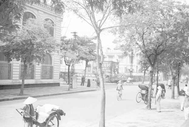 Ein altes Gebäude mit einem Fenster, hinter dem ein weißes Gebäude und Bäume zu sehen sind, mit drei Fahrrädern auf der rechten Seite in der Nähe eines Fußwegs und weiteren Bäumen.