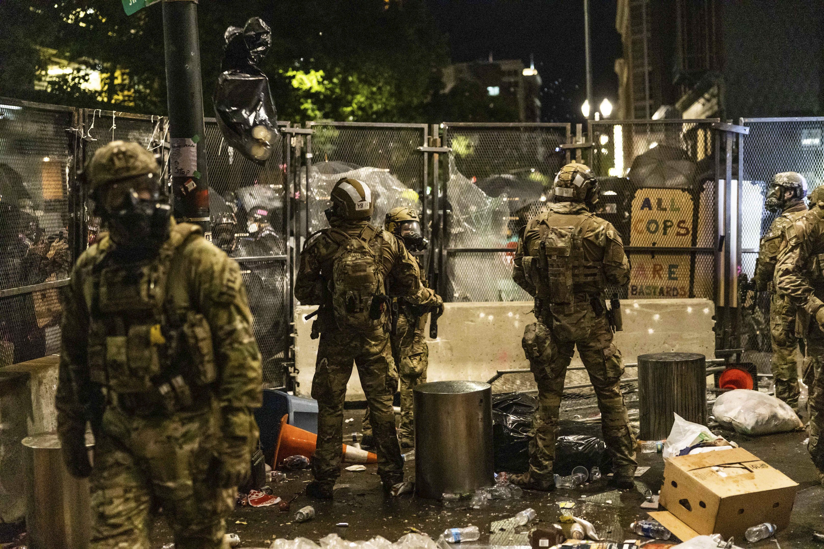 Polizeibeamte in Uniform mit Helmen und Taschen vor einem Zaun bei Nacht stehend, mit Waffen in den Händen, umgeben von Müll und Schutt auf dem Boden und Gebäuden, Bäumen und einem Schild im Hintergrund.