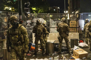 Polizeibeamte in Uniform mit Helmen und Taschen vor einem Zaun bei Nacht stehend, mit Waffen in den Händen, umgeben von Müll und Schutt auf dem Boden und Gebäuden, Bäumen und einem Schild im Hintergrund.
