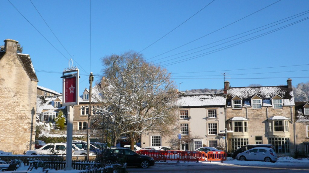 Schneebedeckte Fahrzeuge auf einer Straße mit schneebedeckten Häusern, Bäumen, Polen und Drähten im Hintergrund.