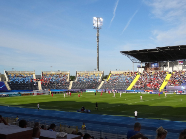 Ein Fussballspiel auf einem grossen Stadionfeld mit Zuschauern auf den Tribünen unter Flutlicht, mit dem Himmel im Hintergrund.