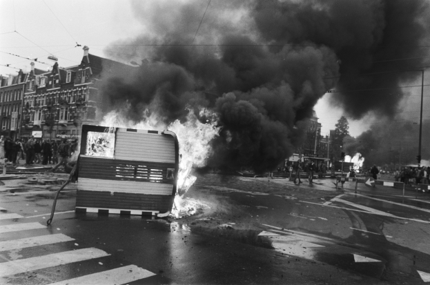 Ein Schwarz-Weiß-Foto eines brennenden Busses am Straßenrand mit Menschen, Masten, einem Schild, Gebäuden und einem bewölkten Himmel im Hintergrund.