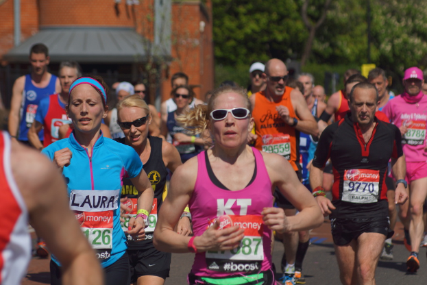 Mehrere Menschen joggen in bunten Shirts, mit einem Geb"ude links und einem Baum rechts.
