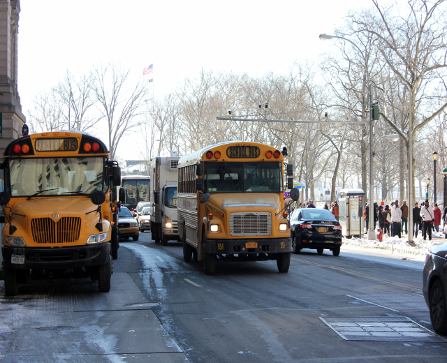 Eine Gruppe von Schulbussen fährt eine Straße mit hohen Gebäuden entlang, Fußgänger auf dem Gehweg, Bäume, Laternenpfähle, eine Fahne und einen sichtbaren Himmel.