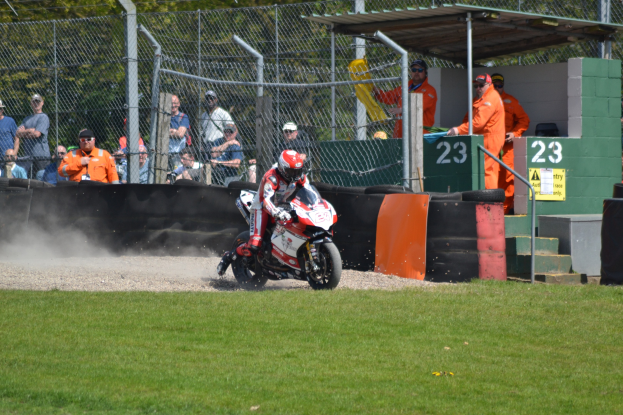 Ein Motorradfahrer mit Helm und Handschuhen fährt über ein grasbewachsenes Feld, mit Zuschauern in Mützen und Schutzbrillen, einem Zaun, einer Treppe mit Geländer und einem numerierten Schuppen im Hintergrund.