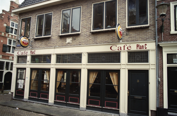 Café-Plan-C-Gebäude in Amsterdam, Niederlande, mit einer Schanktafel, Straßenlaterne, Bäumen und einem bewölkten Himmel.