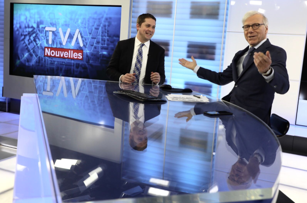 Zwei Männer in Blazern, Krawatten und Brillen sitzen an einem Glastisch in einem Nachrichtenstudio mit einer Tastatur, Maus, Papieren und einem Handy auf dem Tisch und einem Fernseher und Bildschirm im Hintergrund.