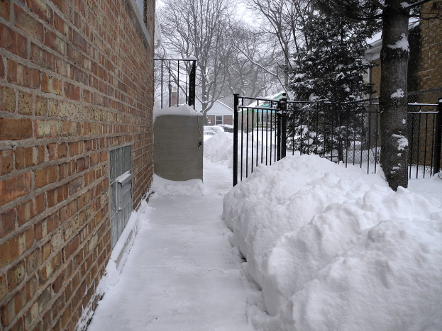 Eine verschneite Szene mit einer Wand links, Schuppen, Bäumen, Gittern und einem Netz im Hintergrund, alles von Schnee bedeckt, mit Schnee am Boden.