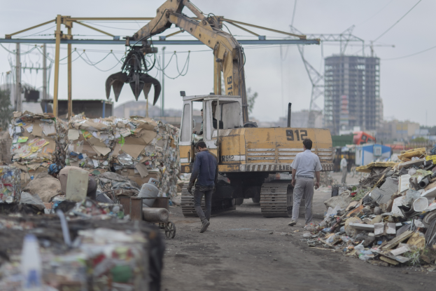 Eine Gruppe von Männern, die eine Schotterstraße entlanggehen, neben einem Haufen Müll mit einem Bagger im Hintergrund, Gebäuden, Bäumen, Polen und Drähten, die in der Gegend verstreut sind.