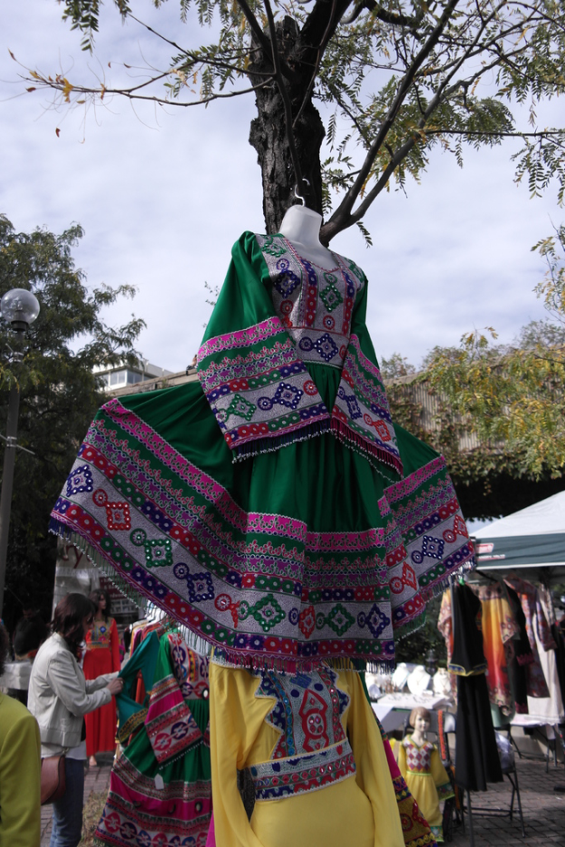 Eine Schaufensterpuppe steht in der Mitte eines belebten Einkaufsgebiets mit verschiedenen Geschäften im Hintergrund, eine Person, die Kleider an einer Stange arrangiert, ein Gebäude und Bäume daneben.