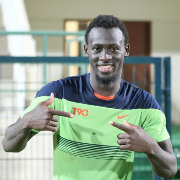 Ein lächelnder Mann in einem grünen und blauen Hemd zeigt auf die Kamera mit einem unscharfen Hintergrund aus einem Geländer, einer Wand und einer Tür.