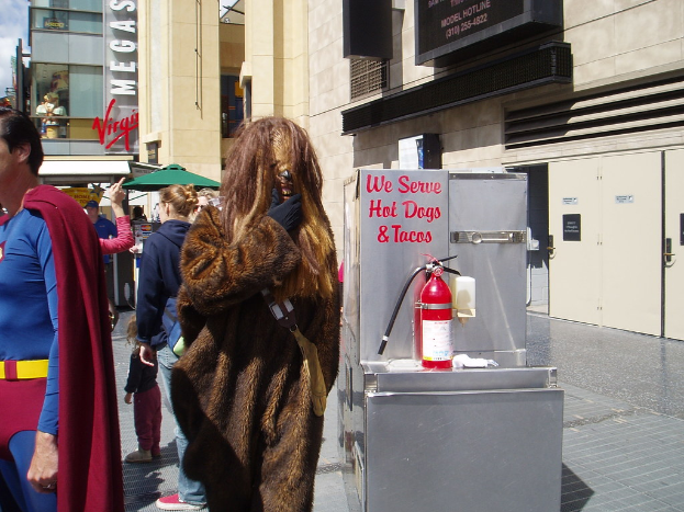 Eine Gruppe von Menschen, einige in Kostümen, versammelt sich vor einem Gebäude mit sichtbarer Beschriftung und Türen, mit einer Box und einem Feuerlöscher in der Nähe.