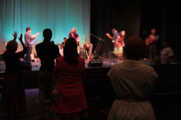 Eine Gruppe von Musikern auf einer Bühne mit Mikrofonen, die Gitarre und Schlagzeug spielen, mit einem klatschenden Publikum vor einem Vorhanghintergrund.
