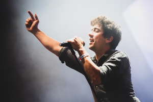 Ein Mann in schwarzem Hemd und Jeans singt in ein Mikrofon auf einer B├╝hne, mit geschlossenen Augen und einer jubelnden Menge im Hintergrund.