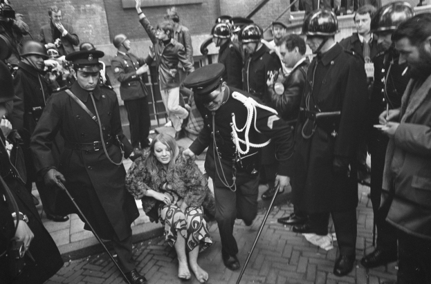 Schwarz-weißes Bild einer Frau, die von der Polizei weggeführt wird, vor einer Menge mit Helmen und Stöcken, mit einem Gebäude mit Treppe und Geländer im Hintergrund.