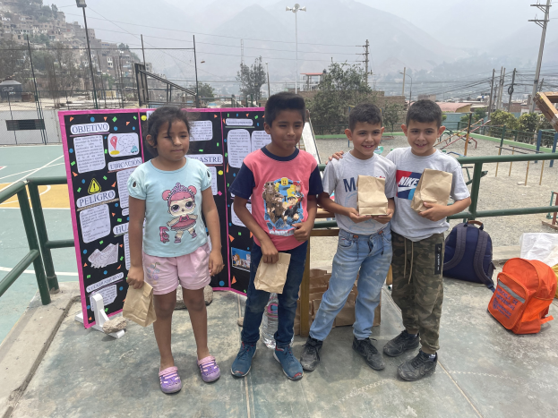 Gruppe von Kindern auf einem Basketballfeld, die Papiertüten halten, mit Tüten und Gegenständen rechts und einer Tafel mit Text links, vor einem Hintergrund aus Zaun, Pfosten, Straßenlaternen, Bäumen, Gebäuden, Hügeln und Himmel.