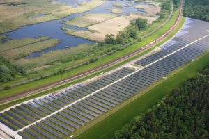 Eine Luftaufnahme eines Solarparks mit zahlreichen Solarpanelen auf einem Feld, umgeben von Bäumen, Gras und Wasser, mit einem Zug, der auf einem nahen Bahnsteig fährt.