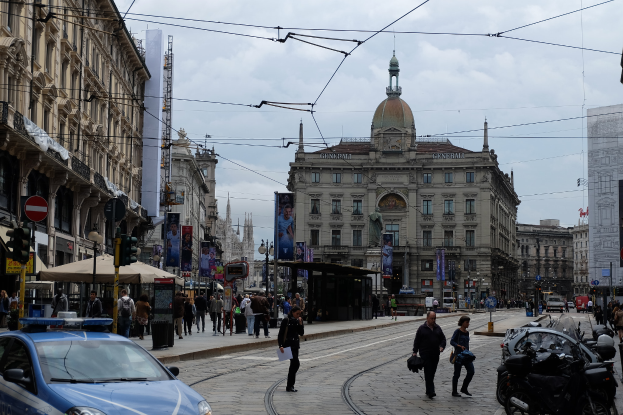 Eine belebte Stadtstraße mit einem geparkten Polizeiwagen, Fußgängern, Fahrzeugen, Gebäuden, Bannern, Laternen und Verkehrsampeln unter einem bewölkten Himmel.
