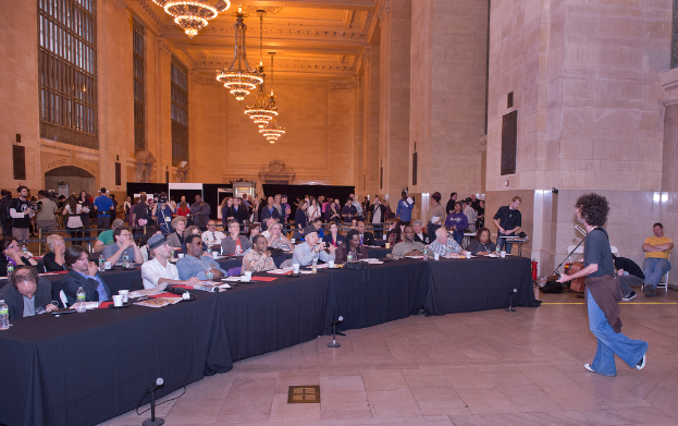 Eine Konzertszene mit Publikum, einem Mann, der auf der rechten Seite Gitarre spielt und singt, einer Wand und Fenstern im Hintergrund, Lichtern oben und einem Boden unten.