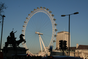 Gebäude und Pfähle rechts, ein großes Riesenrad in der Mitte und eine Skulptur links.