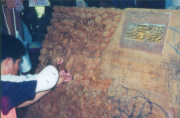 Ein Mann schnitzt auf einer Holztafel mit einem Werkzeug, das Elefanten und Bäume darstellt, mit einem Kalender, einer Tasche und einem Holzpfahl im Hintergrund.