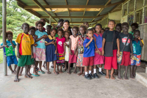 Eine Gruppe von Kindern steht vor einem Gebäude mit einer Frau in der Mitte, Treppen auf der rechten Seite, ein Pfahl auf der linken Seite und einem Hintergrund aus grünem Gras, Bäumen und Himmel.