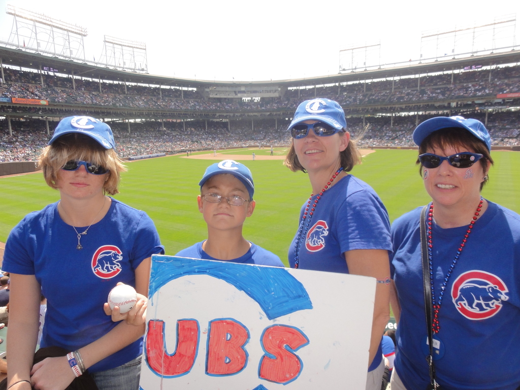 Drei Frauen mit Mützen und Brillen, ein Junge mit Brille und Mütze, der einen Ball und ein Schild hält, in einem Stadion mit Publikum und Feld dahinter, unter einem Himmel mit wenigen Lichtern.