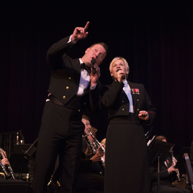 Ein Mann und eine Frau in Fracks, die Mikrofone auf einer Bühne halten, mit einem sitzenden Orchester dahinter in einer dunklen Umgebung.