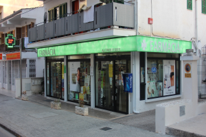 Eine Apotheke an einer Straßenecke mit benachbarten Gebäuden, sichtbaren Geschäftsfronten, Schildern, Versorgungsmasten und -kabeln, fahrenden Fahrzeugen, Topfpflanzen und einem klaren blauen Himmel.