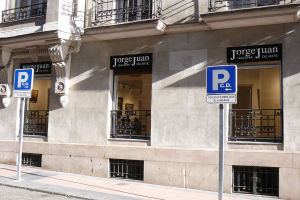 Ein mehrstöckiges Gebäude mit Fenstern, Geländern, Türen und Säulen, das Schilder an Pfosten entlang des Gehwegs und sichtbare Innenobjekte zeigt, die auf eine belebte städtische Umgebung hindeuten.