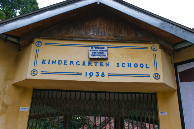 Gelbes Gebäude mit einer "Kindergarten-Schule"-Schilder und einer Wandtafel, einem Gitter unten, Bäumen im Hintergrund und einem klaren blauen Himmel.