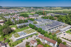 Luftaufnahme eines großen Gebäudes mit Solarmodulen auf dem Dach, umgeben von Häusern, Bäumen, Fahrzeugen auf einer Straße, Hügeln und einem klaren blauen Himmel, identifiziert als Hauptsitz eines deutschen Solarenergieunternehmens.