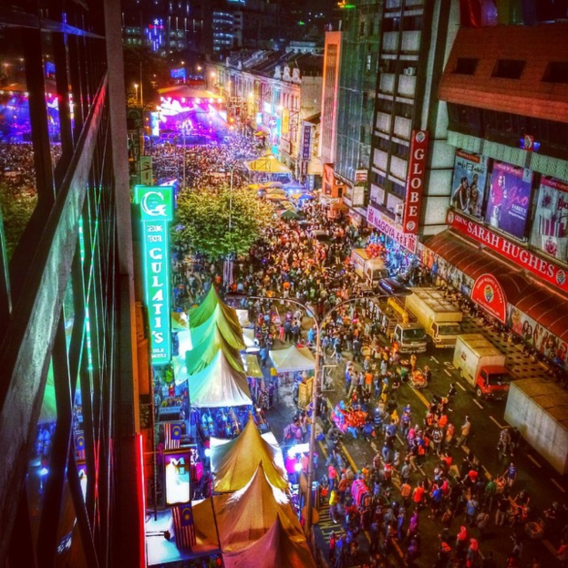 Volles Karnevalstreiben mit Zelten, Buden, Gebäuden und Bannern, mit beleuchteter Fläche im Hintergrund.