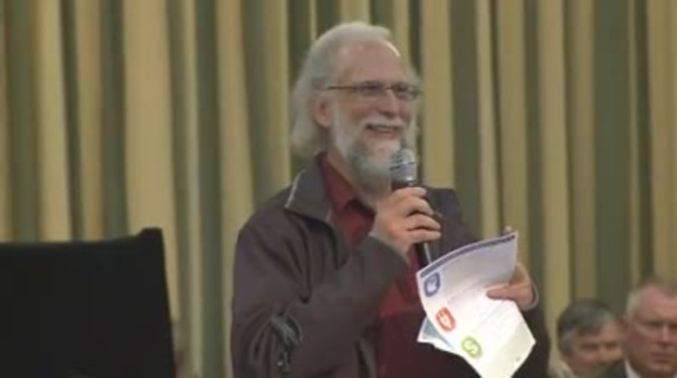 Eine Person steht an einem Podium und hält ein Mikrofon und Papier in der Hand, mit Stuhlpersonen und einem Vorhang im Hintergrund.