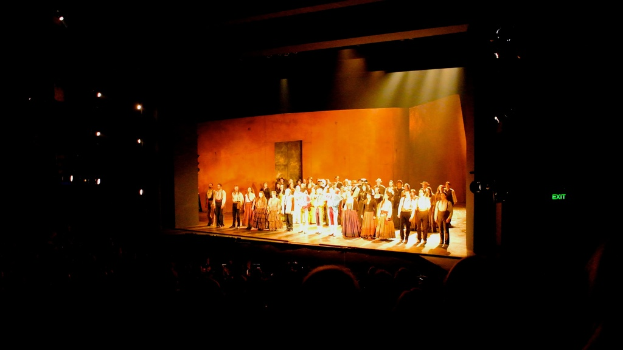 Eine Bühne mit Menschen in altertümlicher Kleidung, die von einem Publikum und Scheinwerfern links umgeben sind.