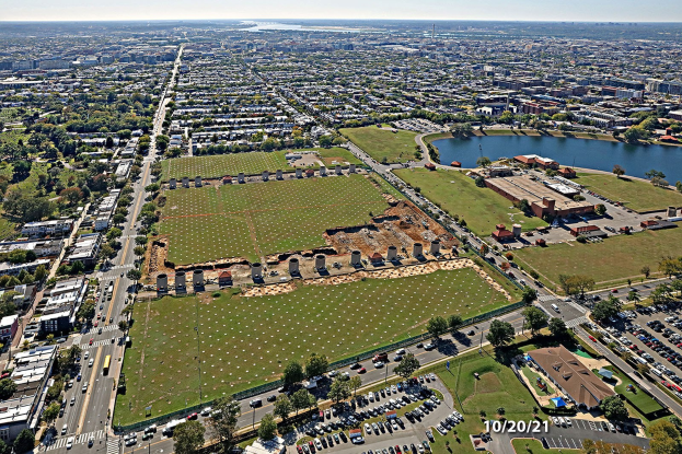 Luftaufnahme einer Stadt mit Gebäuden, Bäumen, Gras, Straßen mit Fahrzeugen, Wasser und Wolken, die einen großen zentralen Platz für einen Park vorschlagen.