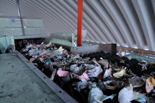 Ein großer Haufen Müll auf einem Förderband in einer Recyclinganlage, mit einer Maschine im Vordergrund und einem Metalldach darüber.