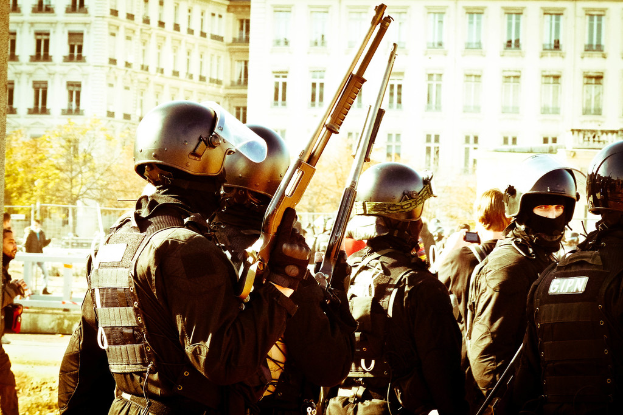 Mehrere Personen in Uniform und mit Helmen stehen und halten Gewehre, im Hintergrund sind Straßen, Bäume, Gebäude und Fenster sichtbar.