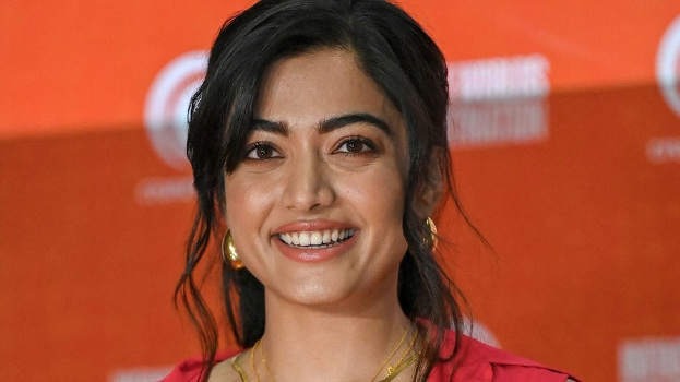 Eine Frau in einem roten Kleid mit einem eleganten Dutt und dezentem Make-up steht lächelnd da, trägt schwarze High Heels und hat ein hellgelbes Banner mit der Aufschrift 'Sonam Kapoors Beste Schönheits Looks vom Cannes Film Festival' im Hintergrund.
