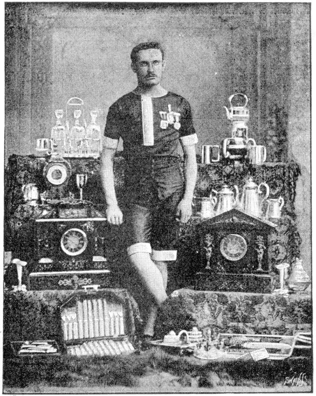 Schwarzes und weißes Foto eines Mannes in einem Fußballtrikot, der vor einem Tisch mit Pokale, Trophäen und anderen Gegenständen steht, beschriftet mit "1909-1910 WM-Gewinner."