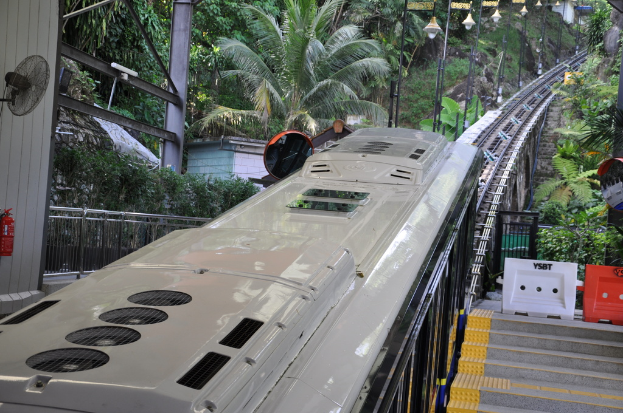 Ein Zug auf Eisenbahnschienen mit Gebäuden, Polen, einem Ventilator, einem Feuerlöscher und einem Geländer auf der linken Seite, verschiedenen Gegenständen und einer Treppe auf der rechten Seite und Bäumen im Hintergrund.