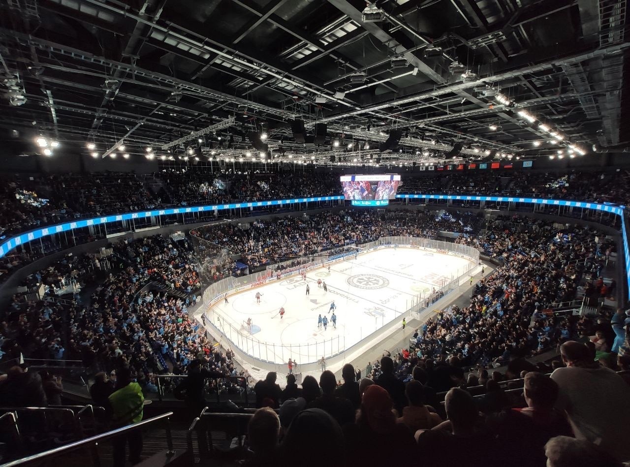 Ein Eishockeyspiel wird im O2 Arena in London gespielt, mit Zuschauern auf den Tribünen und Spielern auf dem Eis, unter hellen Scheinwerfern mit einem Bildschirm an der Spitze des Bildes.